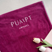 Fuchsia gym towel with 'Pumpt Apparel' branding, a bottle, and tulips on a light background