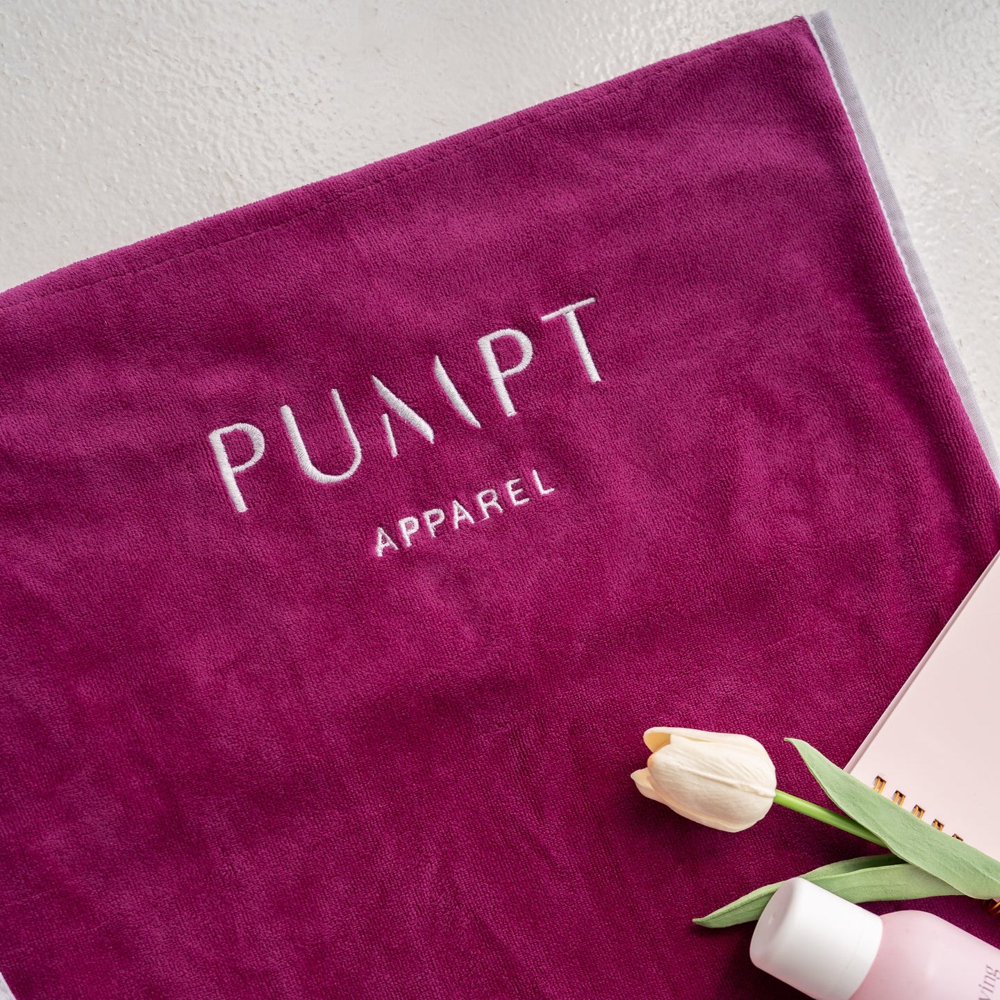 Fuchsia gym towel with 'Pumpt Apparel' branding, a bottle, and tulips on a light background