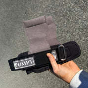 A hand holding a black and grey adjustable wrist strap with a grey fabric cuff, featuring a white label that reads 'PUMPT'. The background is a textured gray asphalt surface.