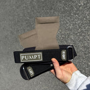 A person's hand holds a pair of wrist wraps, black and khaki with a black adjustable strap, against an outdoor asphalt background. The straps feature white labels reading 'LEFT' and 'RIGHT' and a central label reading 'PUMPT'.