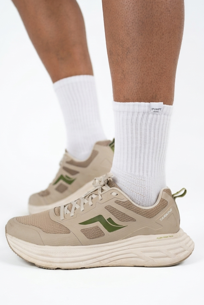 Beige athletic sneakers with green accents worn with white crew socks, shown on a model's feet against a white studio background.
