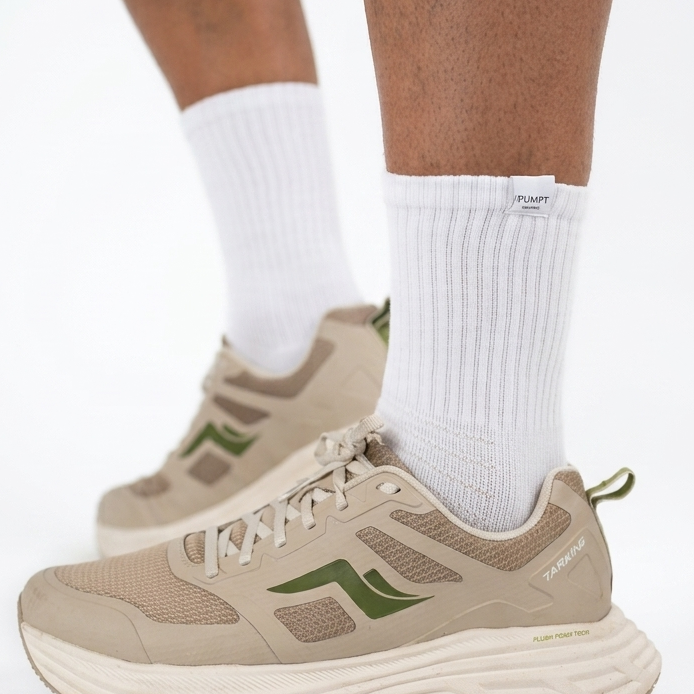 Beige athletic sneakers with green accents worn with white crew socks, shown on a model's feet against a white studio background.
